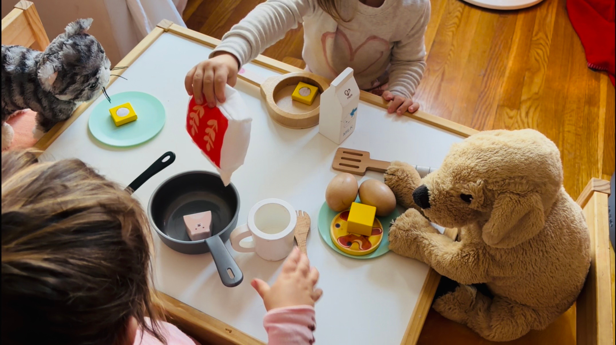 Toddlers engaged in pretend play kitchen activity with wooden toy food, pan, milk bottle, and stuffed animals, supporting imaginative play and early childhood homeschool learning at home