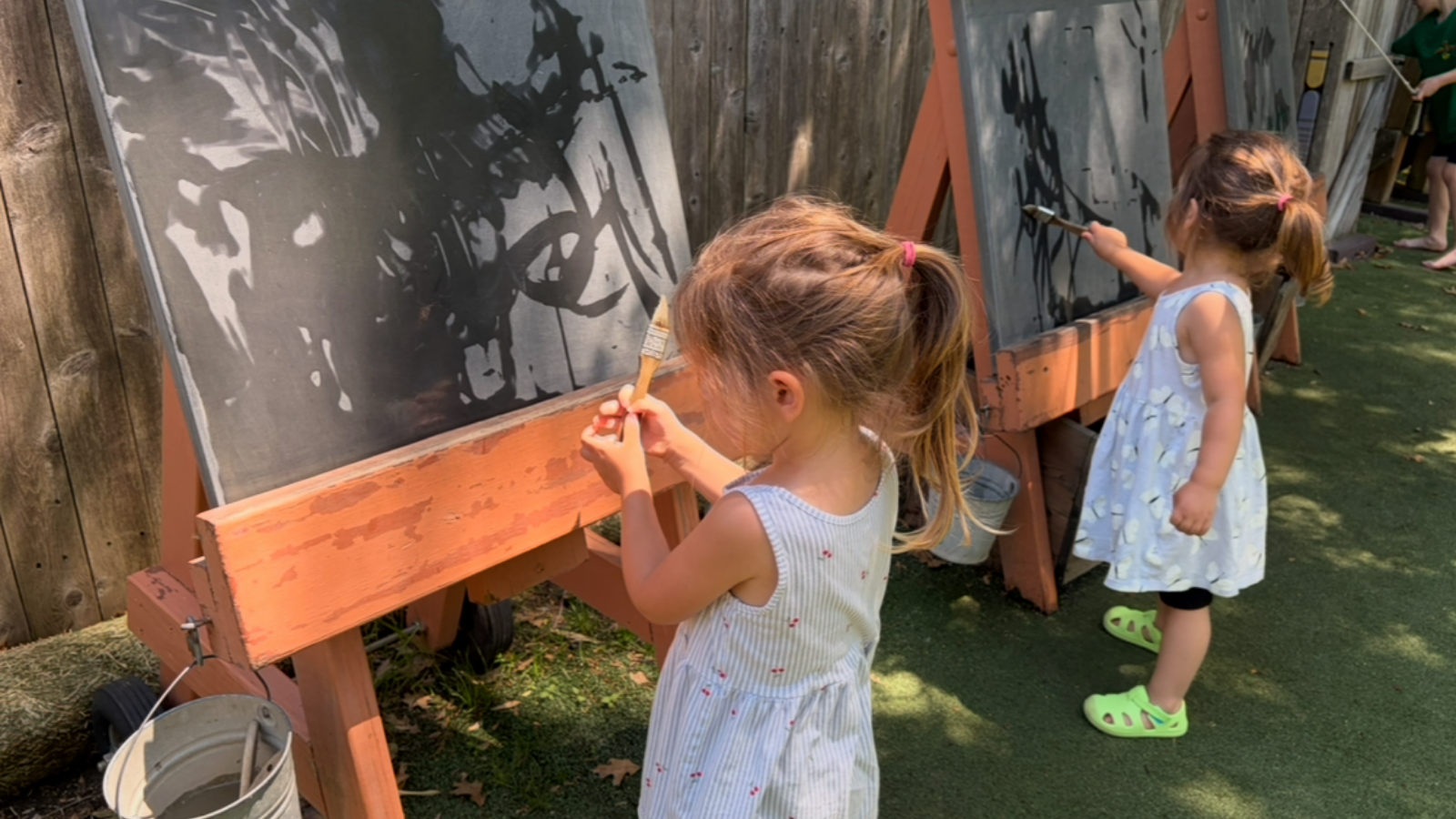 Young children painting outdoors at a large chalkboard during open-ended play.