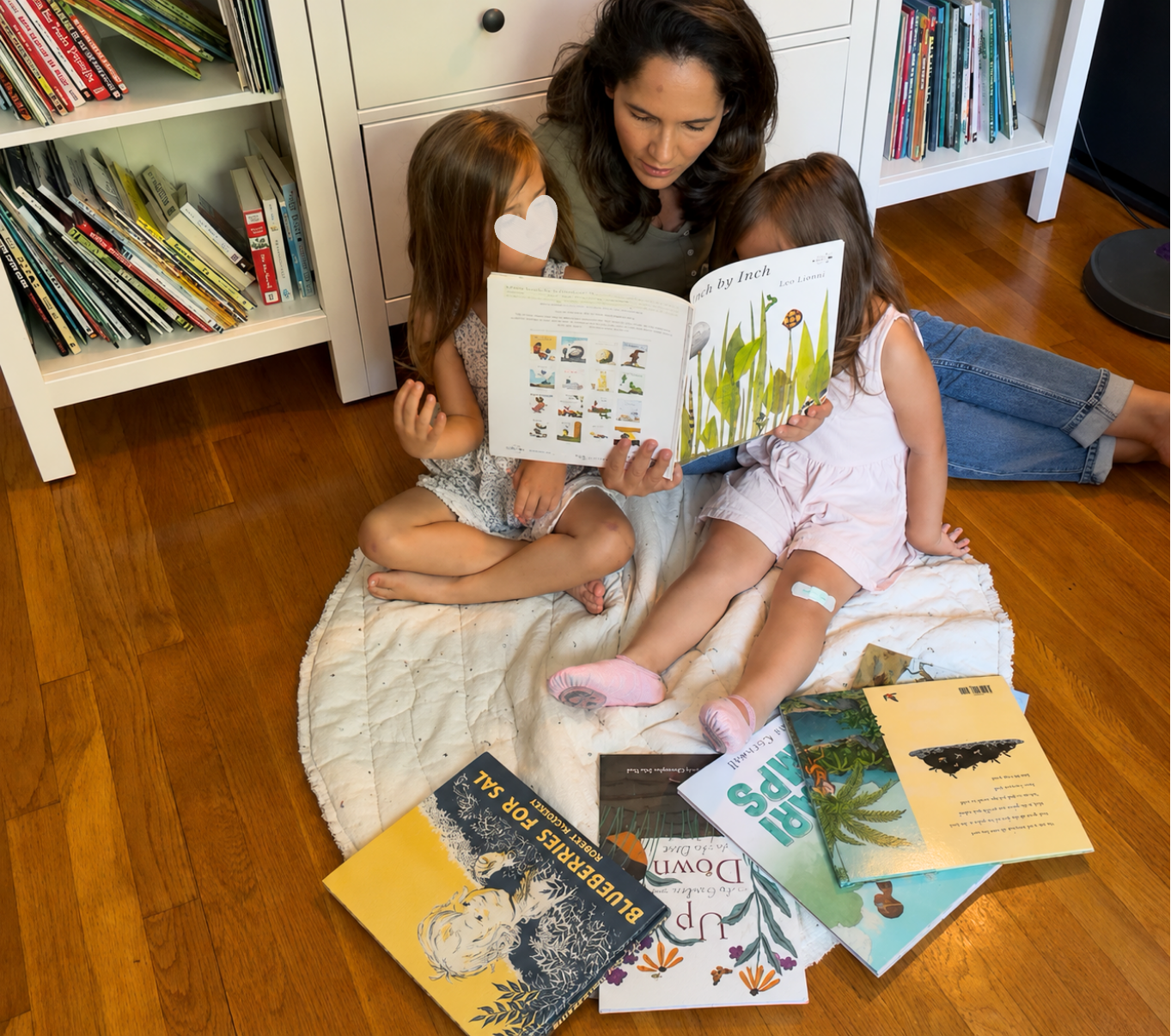 A mother reads a picture book with her two young daughters while surrounded by a collection of children's books, creating a cozy story-led learning moment at home.