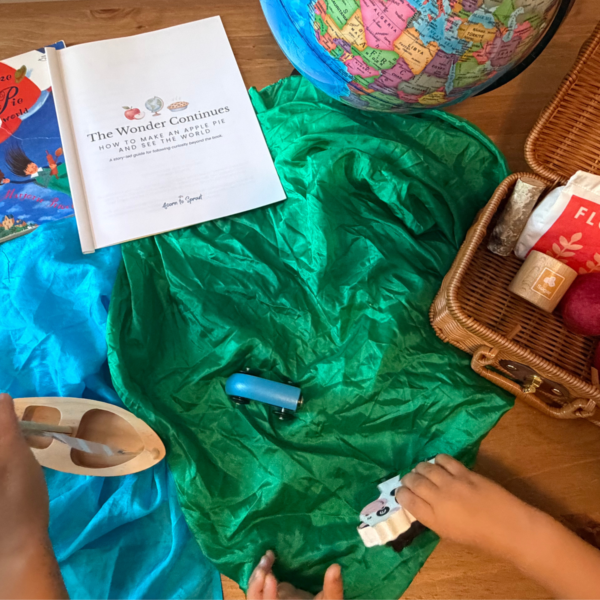 Around-the-world pretend play with a globe, play silks, suitcase, toy vehicles, and The Wonder Continues Apple Pie preschool guide