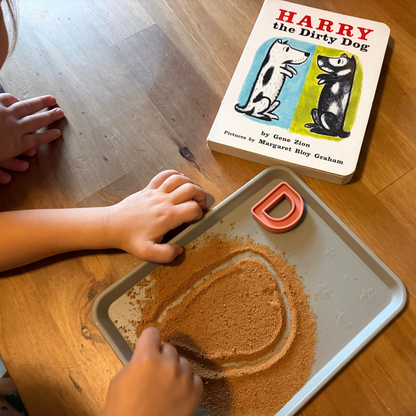 Child tracing the letter D in a sensory tray beside Harry the Dirty Dog.