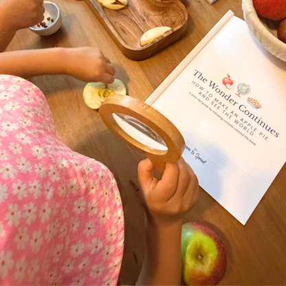 Child examining an apple with a magnifying glass beside The Wonder Continues How to Make an Apple Pie and See the World guide
