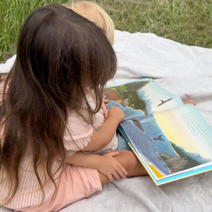 Young children reading The Snail and the Whale together outdoors on a blanket.