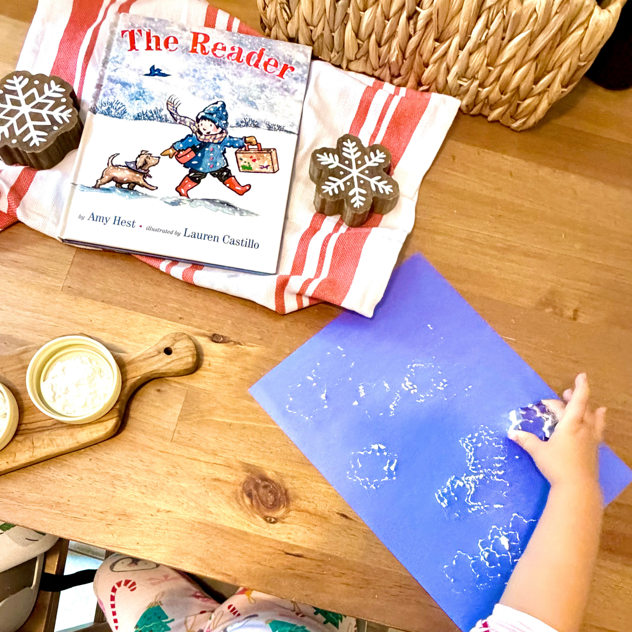Child creating snowflake art at the table beside The Reader by Amy Hest.