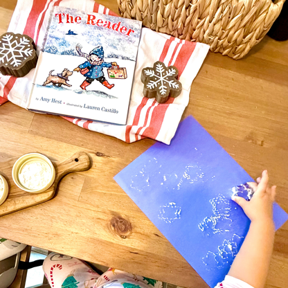 Child creating snowflake art at the table beside The Reader by Amy Hest.